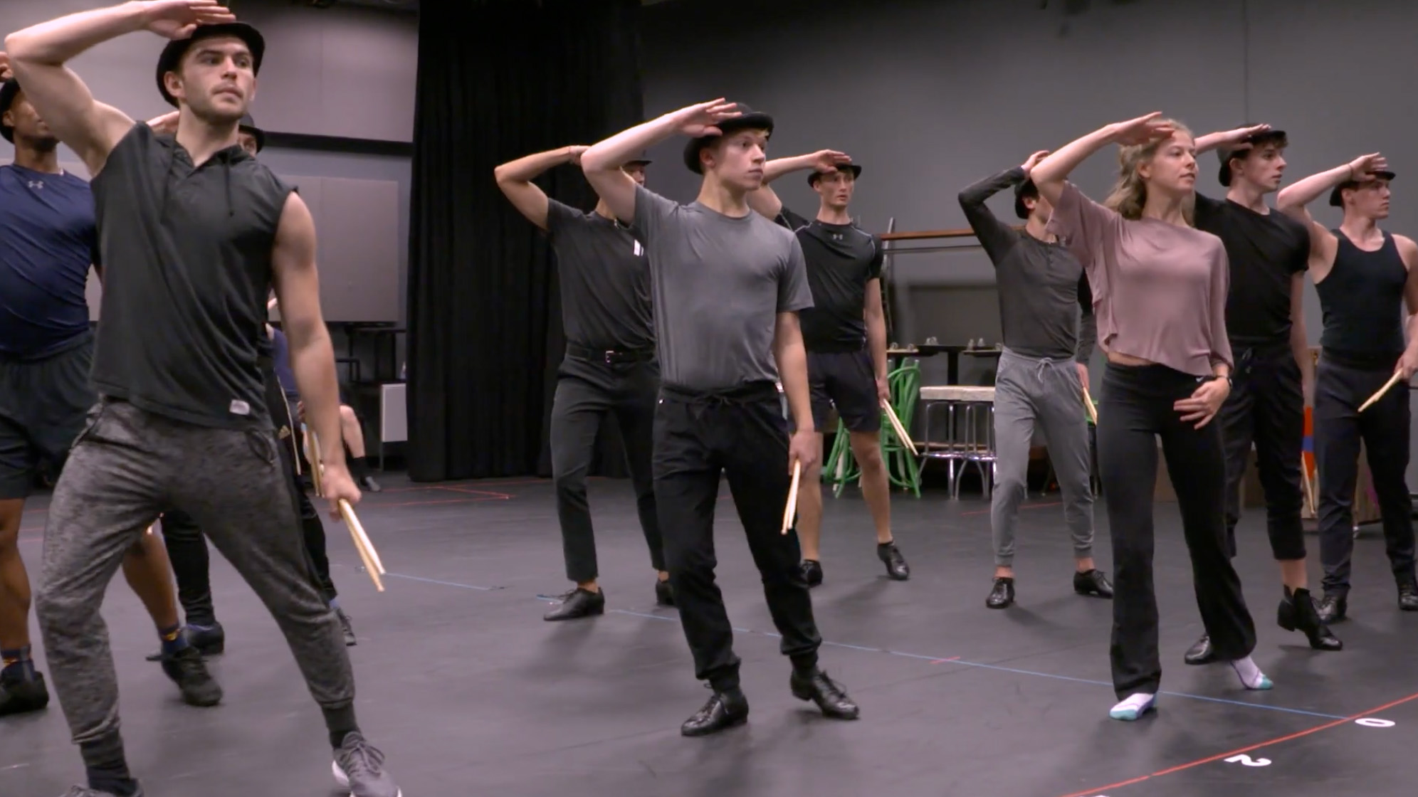 A group of dancers stand in a studio, posing with arms raised and holding batons.