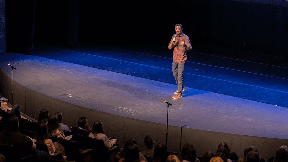 A speaker stands center stage in front of an audience during the Flint Future Teachers Academy in April 2025.