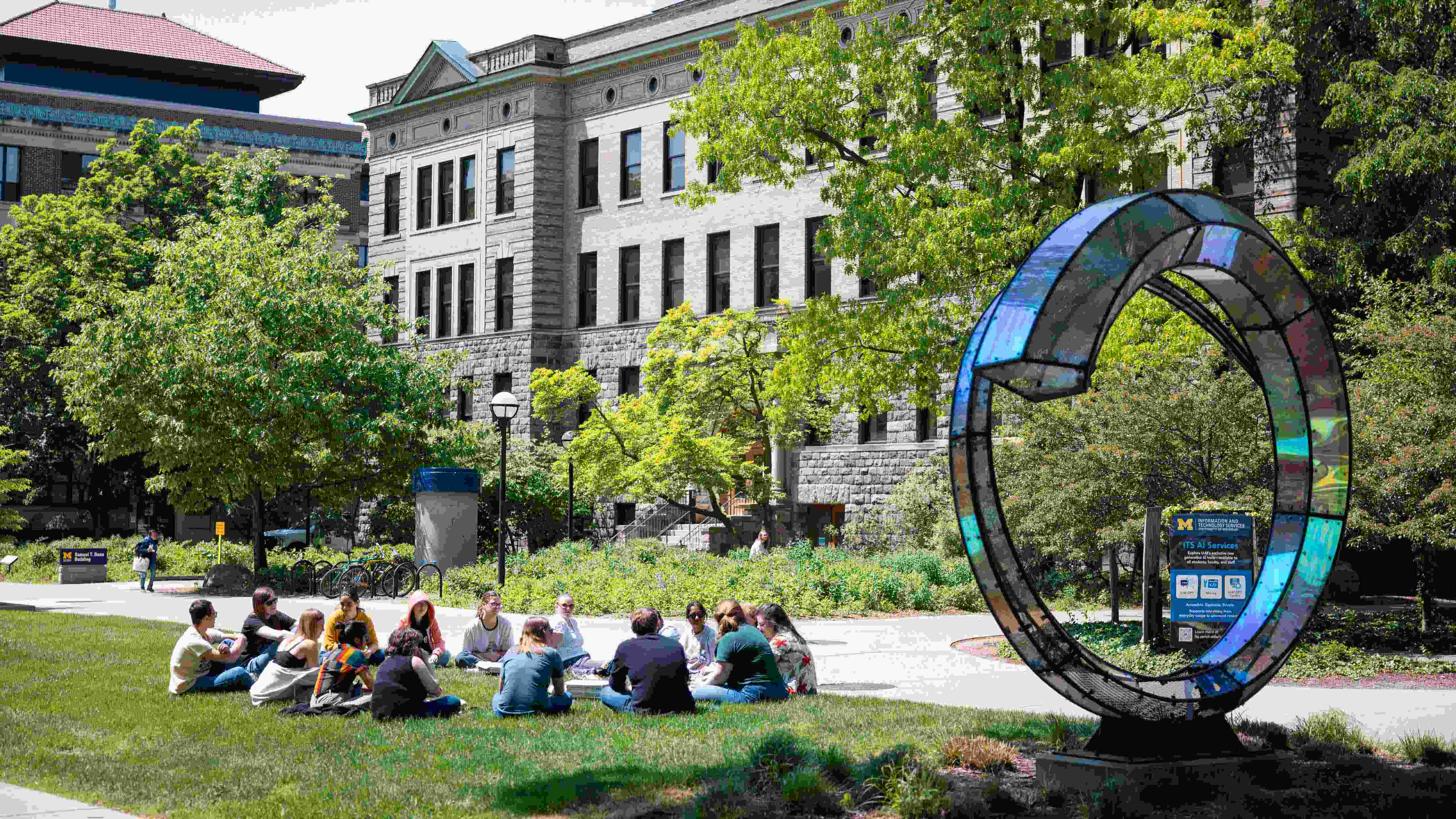 A group of students sitting in a circle on the ground outside. There’s a circular sculpture to the right of them and a building behind them.