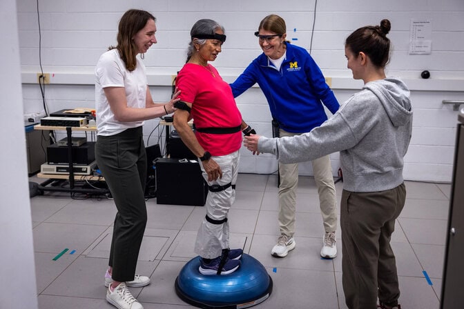 Geeta Peethambaran stands on a balance trainer, testing steadiness while 3 researchers stand close by to help if she loses her balance.