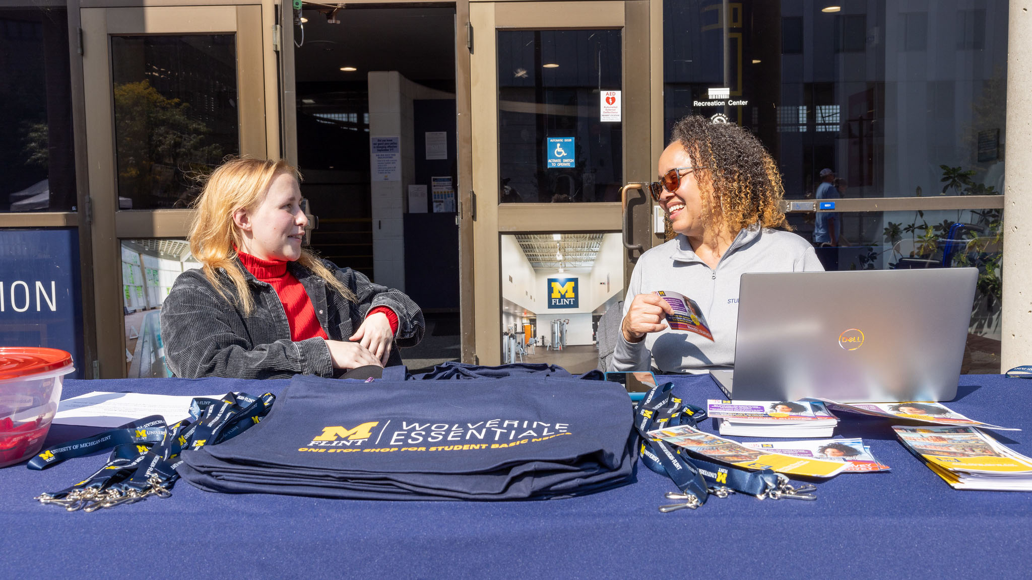 Two people sit at a table covered with University of Michigan materials, including brochures, lanyards, and tote bags, in front of a building entrance with glass doors.