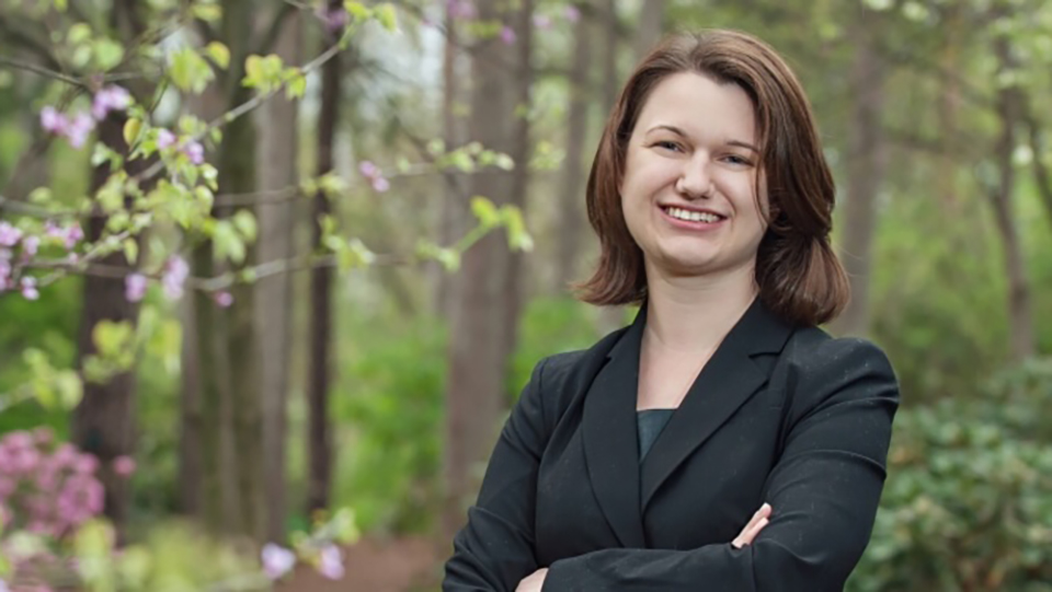 A woman standing outside with her arms crossed and smiling with woods and flowers in the background. 