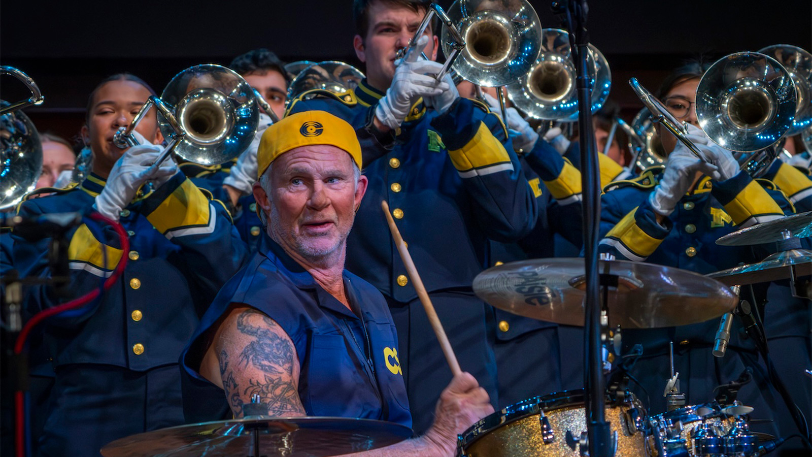 Chad Smith sitting at a drum kit in front of trumpet players, all dressed in maize and blue