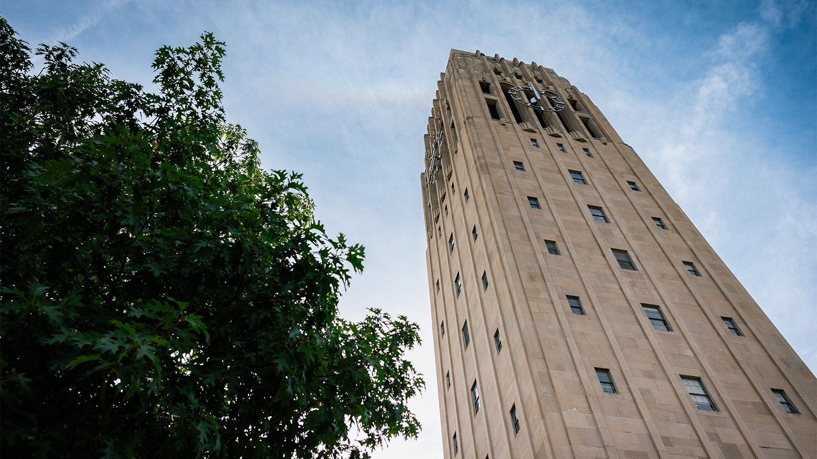 The Burton Memorial Tower, with its stone exterior and large clocks, extends skyward next to lush green foliage under a bright, partly cloudy sky.