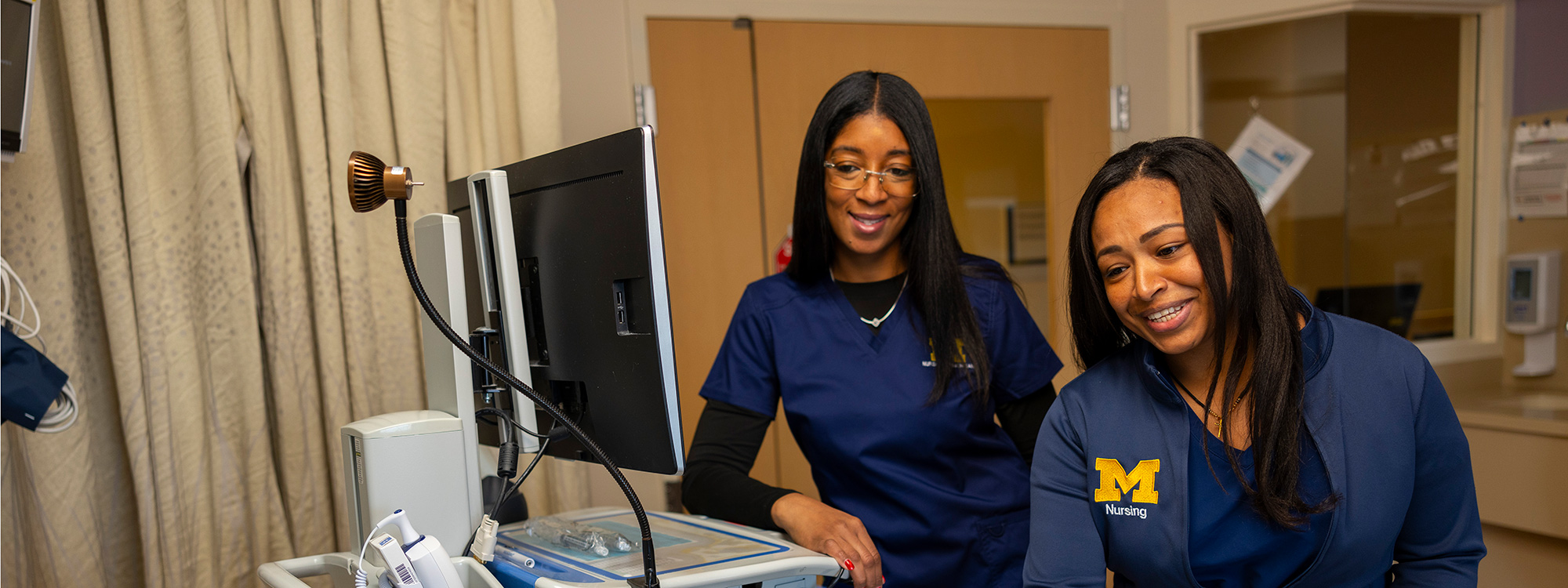 Two University of Michigan nursing professionals smile while looking at a medical monitor in a clinical setting.