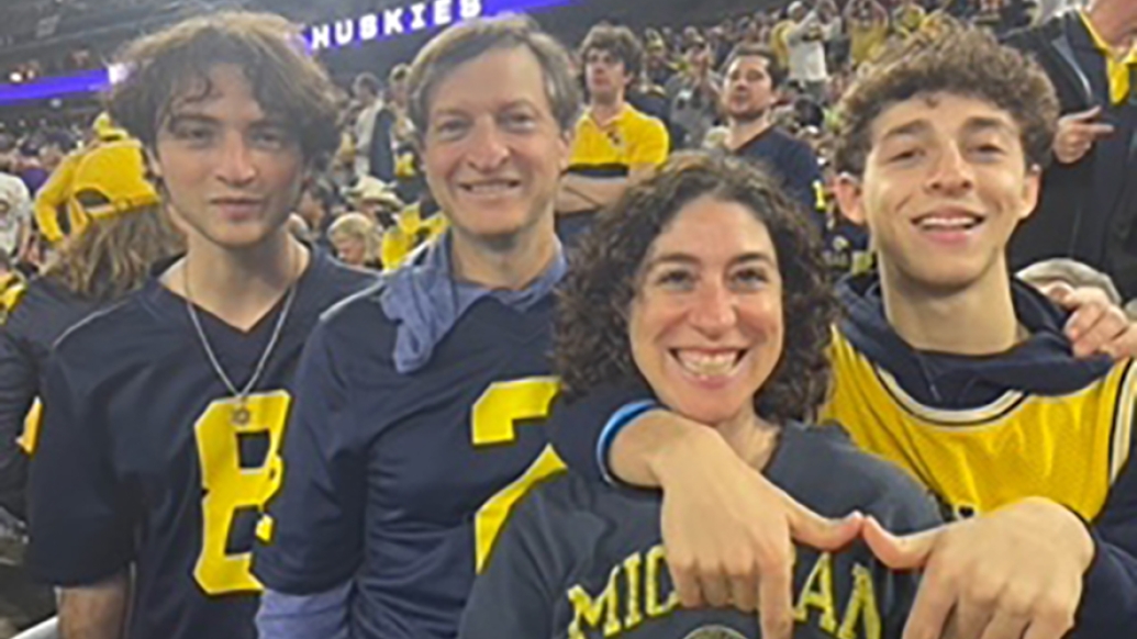 Four people stand together at a packed stadium wearing Michigan shirts.