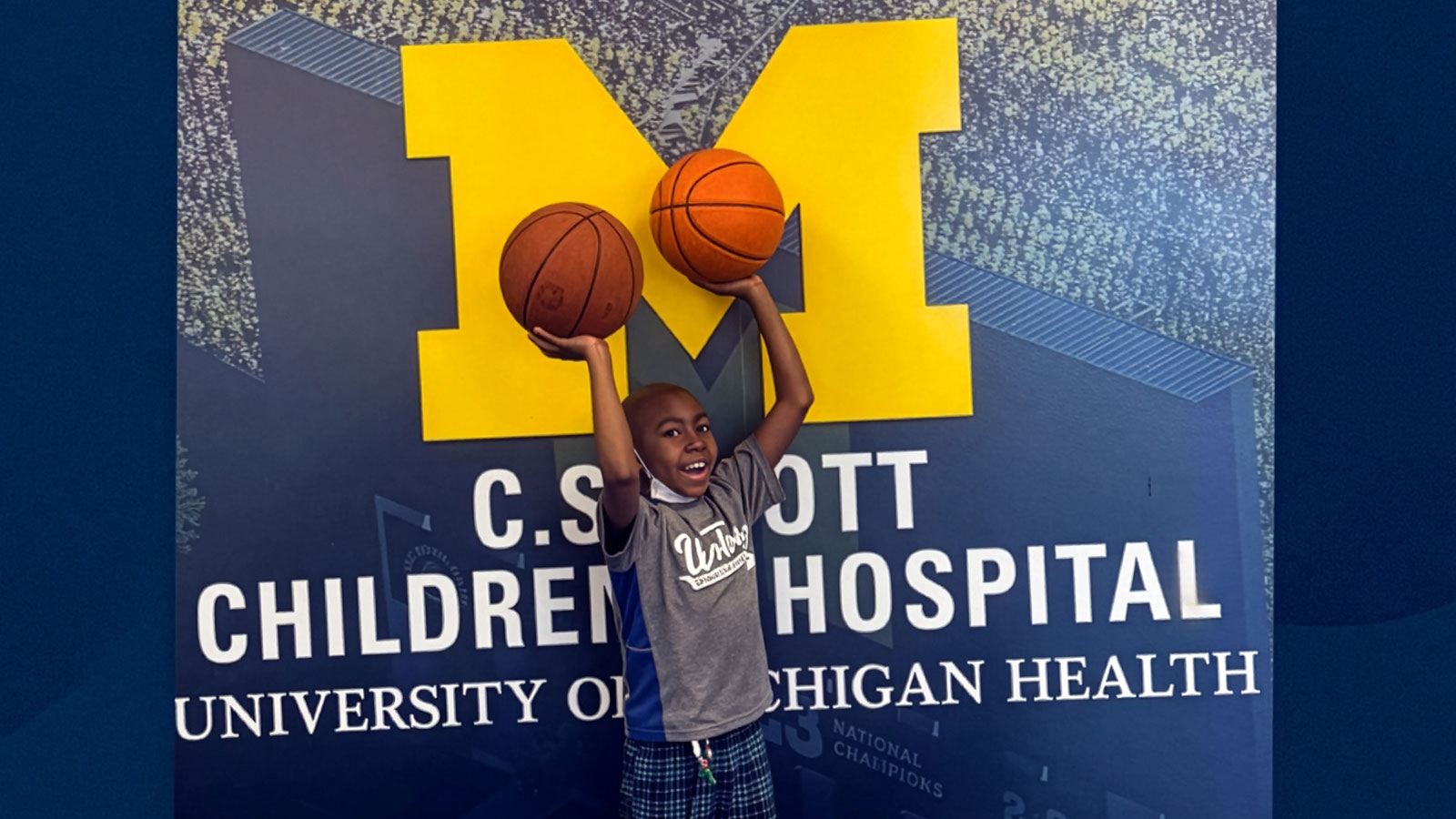 Elijah Dottery smiling while holding two basketballs in front of a University of Michigan mural.