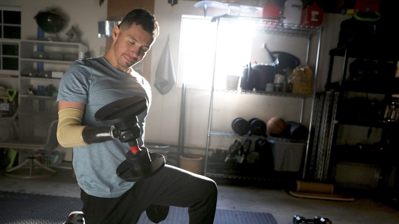 A person wearing a light blue t-shirt kneels on one knee in a home gym or garage while lifting a dumbbell with a prosthetic arm.