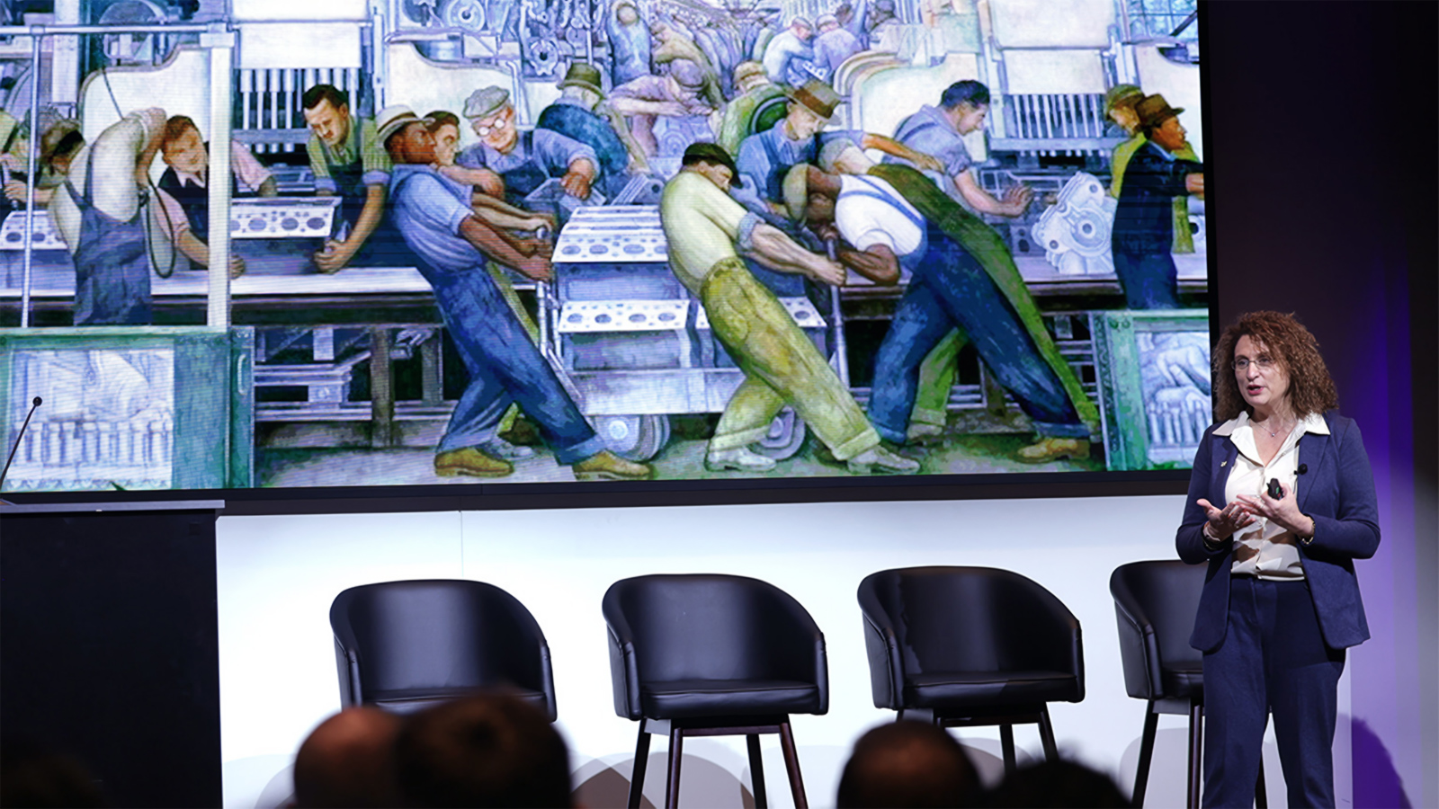 Engineering professor Valeria Bertacco speaks in front of a screen displaying Diego Rivera’s “Detroit Industry” mural at Newlab in Detroit during the inaugural Automotive Semiconductor Forum.