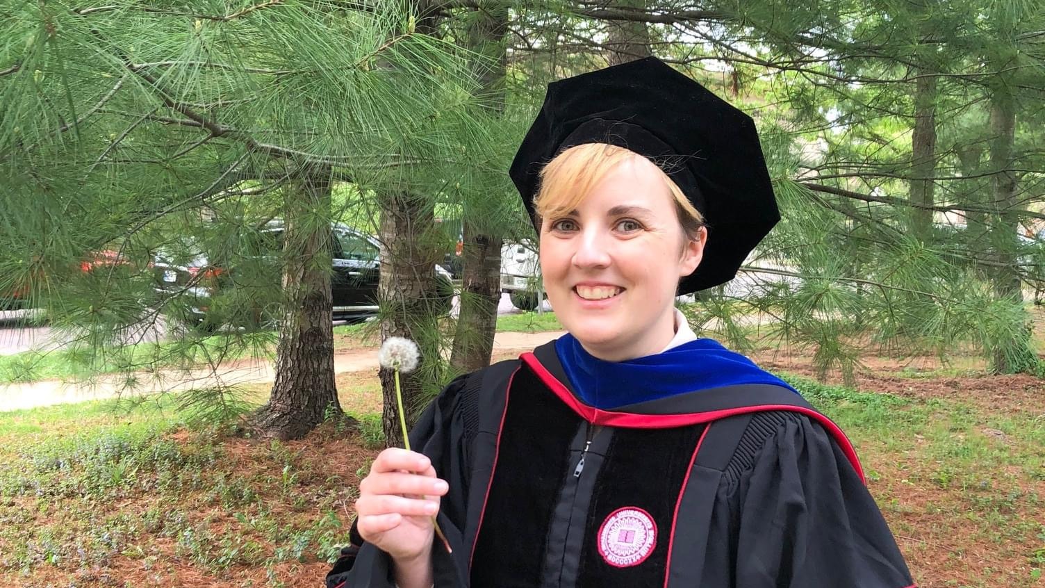 Maggie Rossman posing with a dandelion in graduation cap and gown