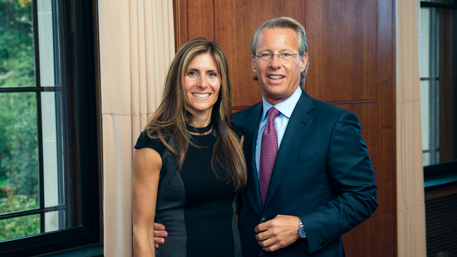 Nate and Catherine Forbes stand side by side, dressed formally, inside a room with wood panels and large windows.
