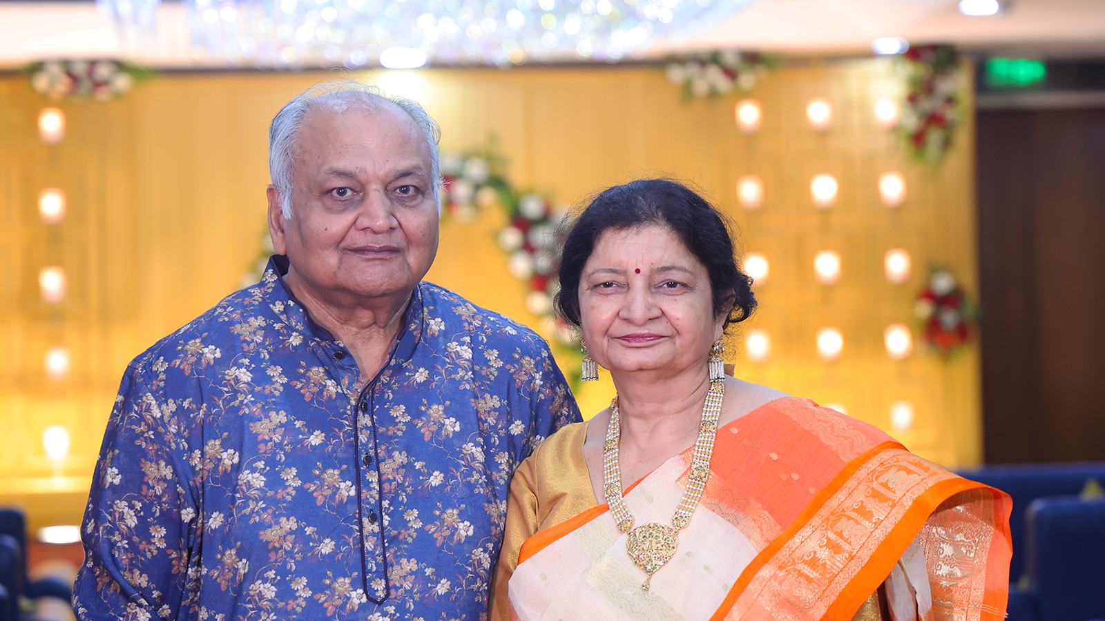 Ashok and Sarita Jain standing together at an event, wearing traditional clothes, with a bright, decorated background.