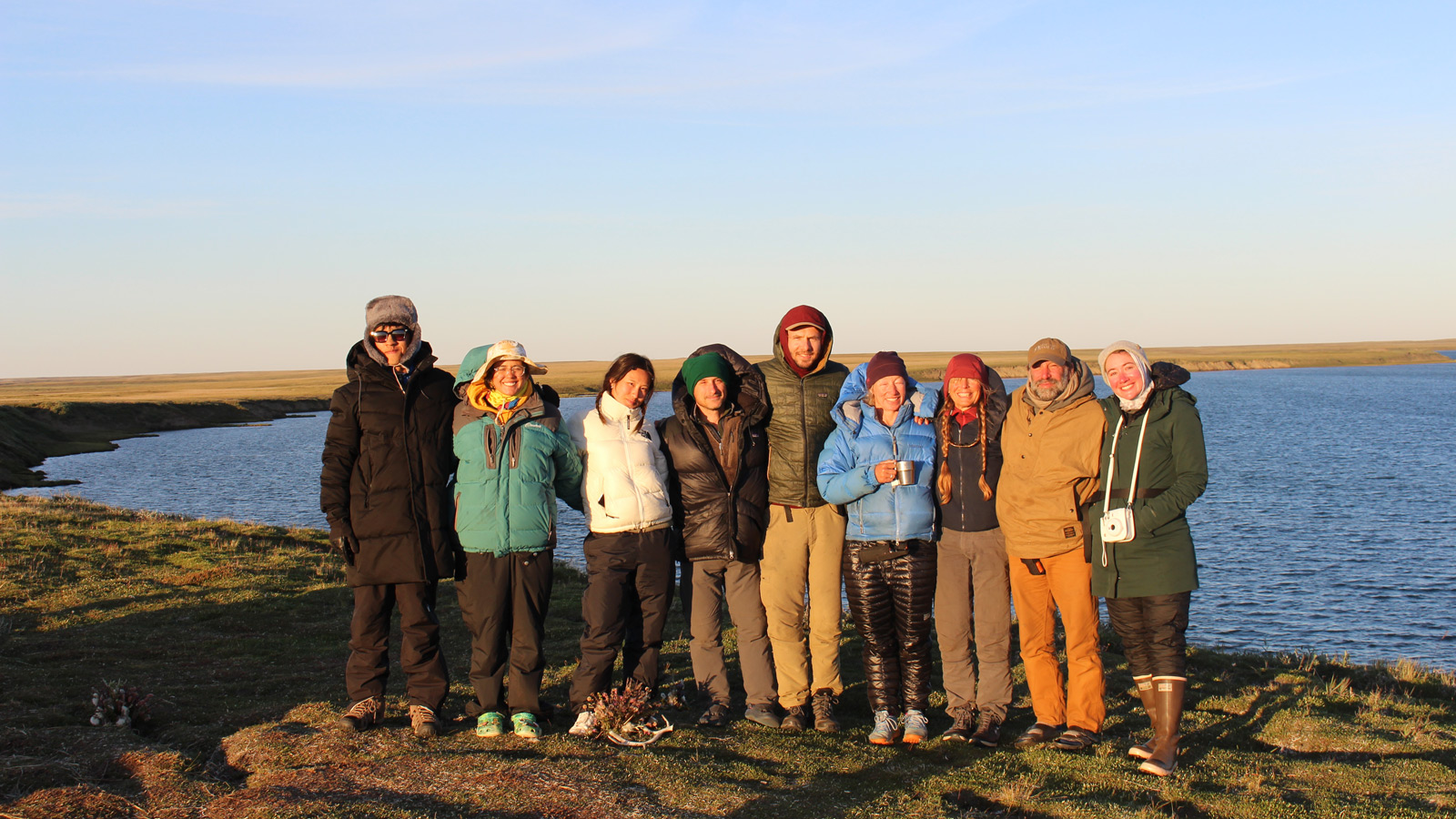 Ten members of SEAS in insulated jackets and boots pose arm-in-arm on grassy ground next to a calm, winding river, with expansive plains and a blue sky behind them.