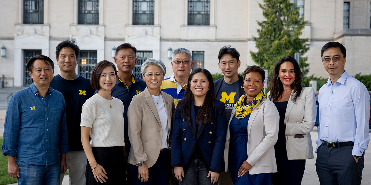 Eleven people standing together outside a university building, some in University of Michigan shirts and accessories.