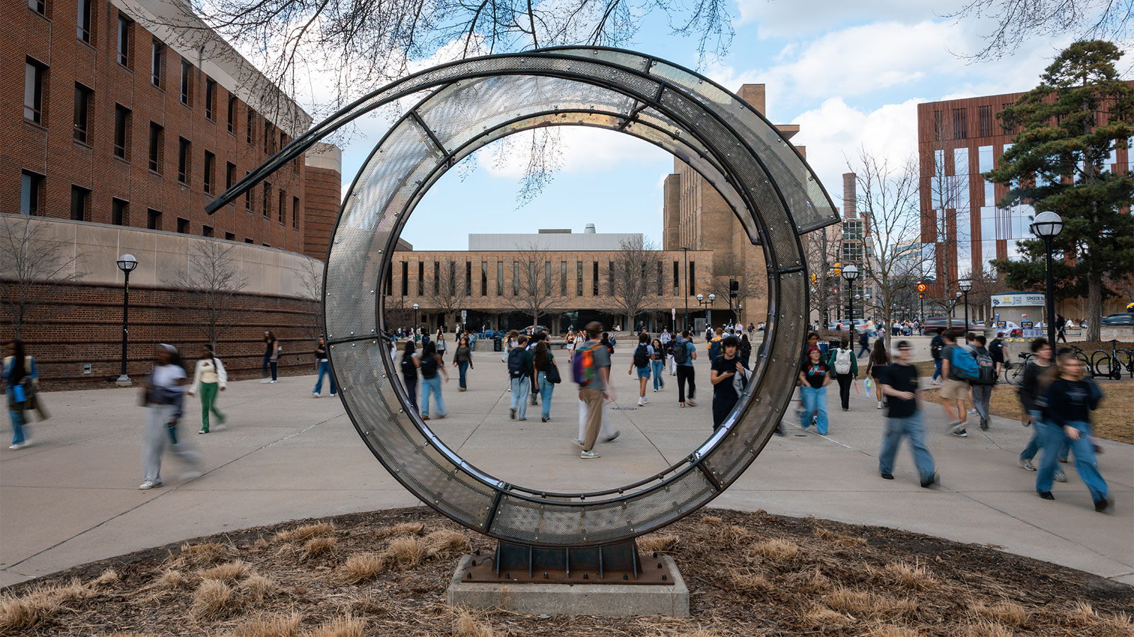 A group of students walks across a plaza at the University of Michigan. In the foreground stands a large, circular metal mesh sculpture, framing a view of campus buildings under a partly cloudy sky.