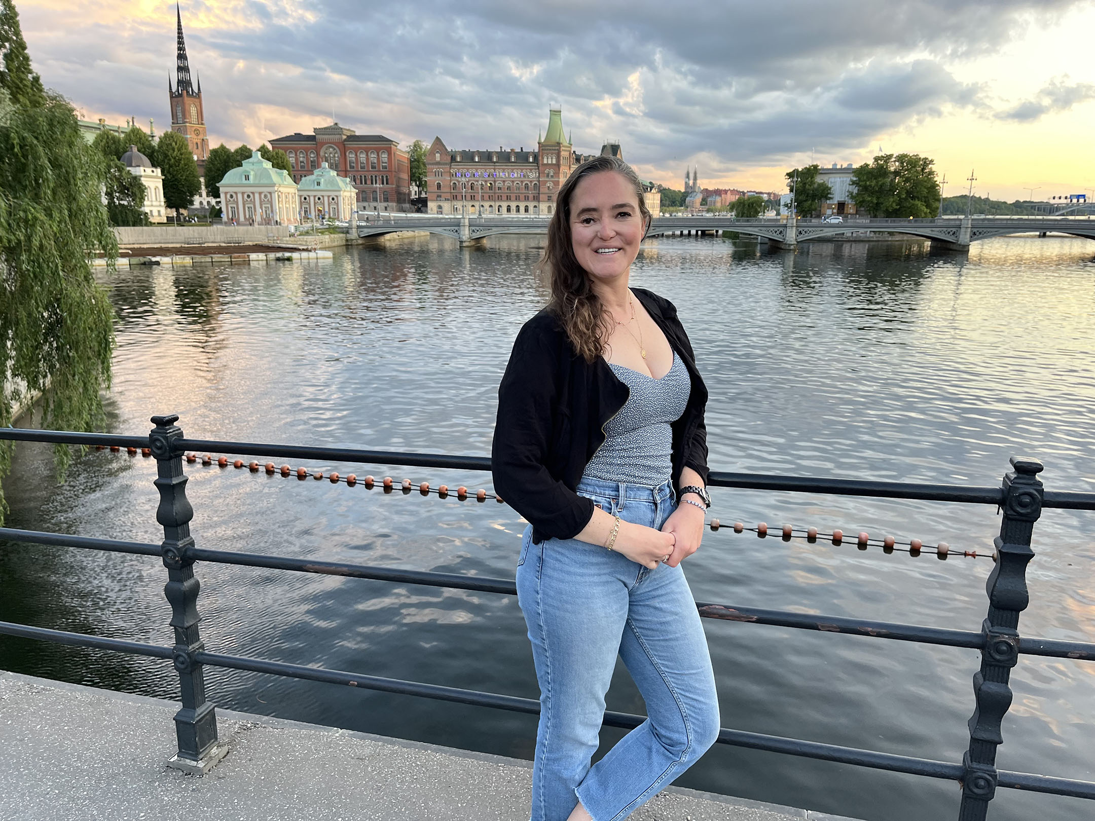 Laura Raines leans on a wrought iron railing alongside a canal in Stockholm, Sweden. Historic buildings and a church steeple are visible on the other side of the canal.