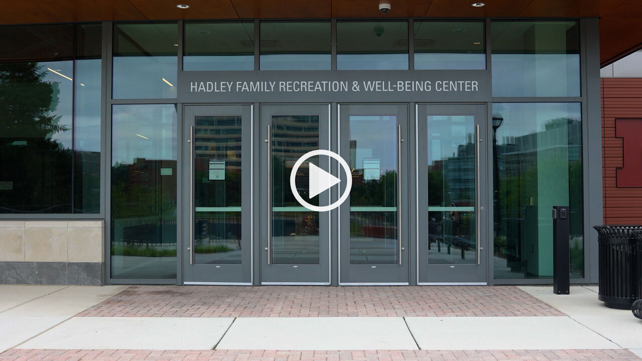 Glass-front entrance to the Hadley Family Recreation & Well-Being Center, with four gray-framed doors beneath the building’s name. A play button linking to a video is overlayed.