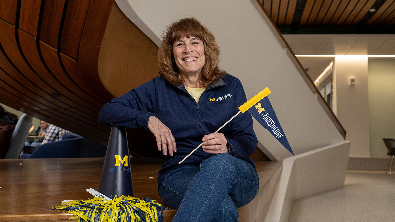 Pat sits on a curved wooden bench in a modern building, smiling and holding a University of Michigan School of Kinesiology pennant.
