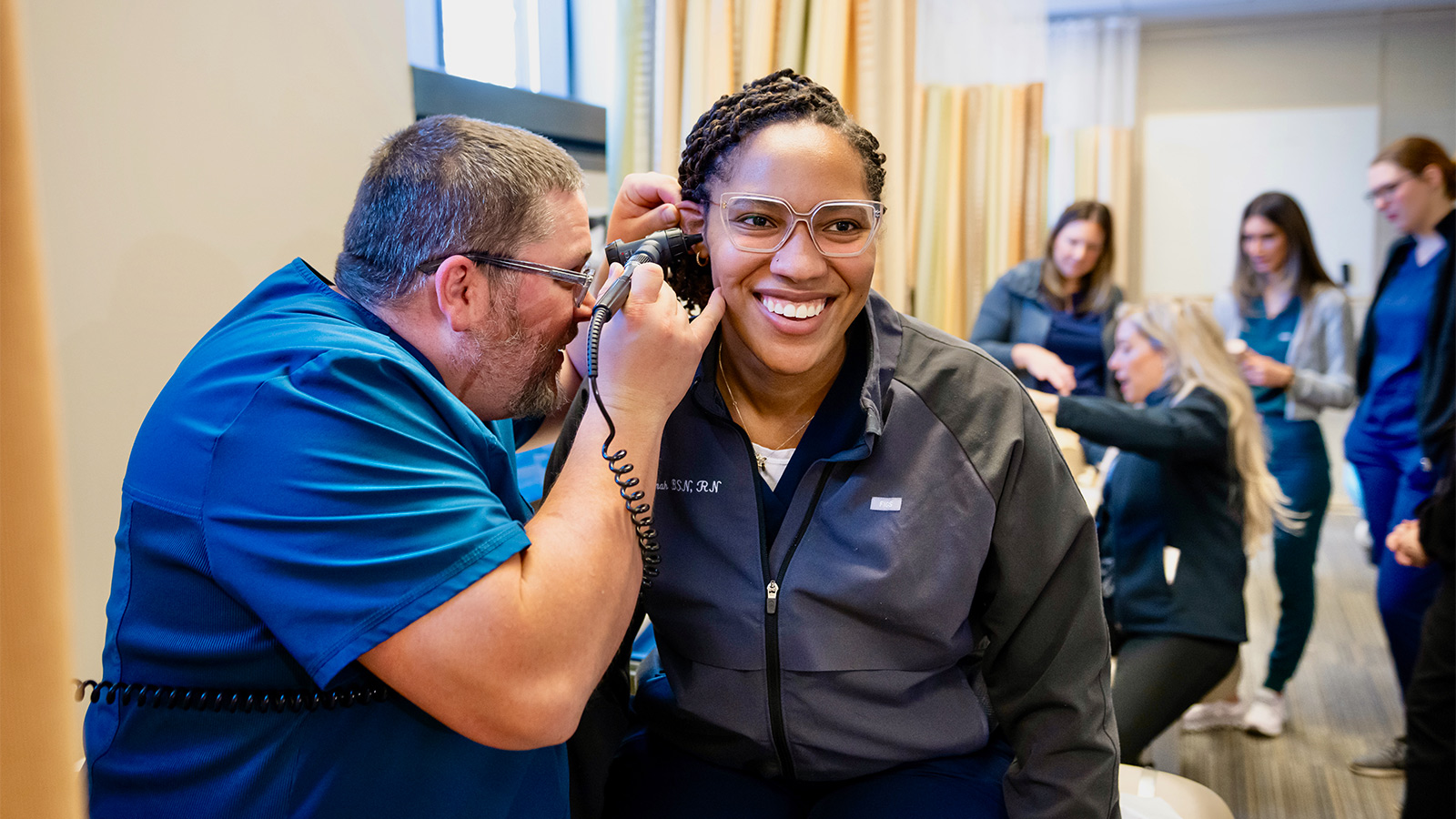 A medical professional uses an otoscope to examine the ear of a smiling patient in a clinic setting.