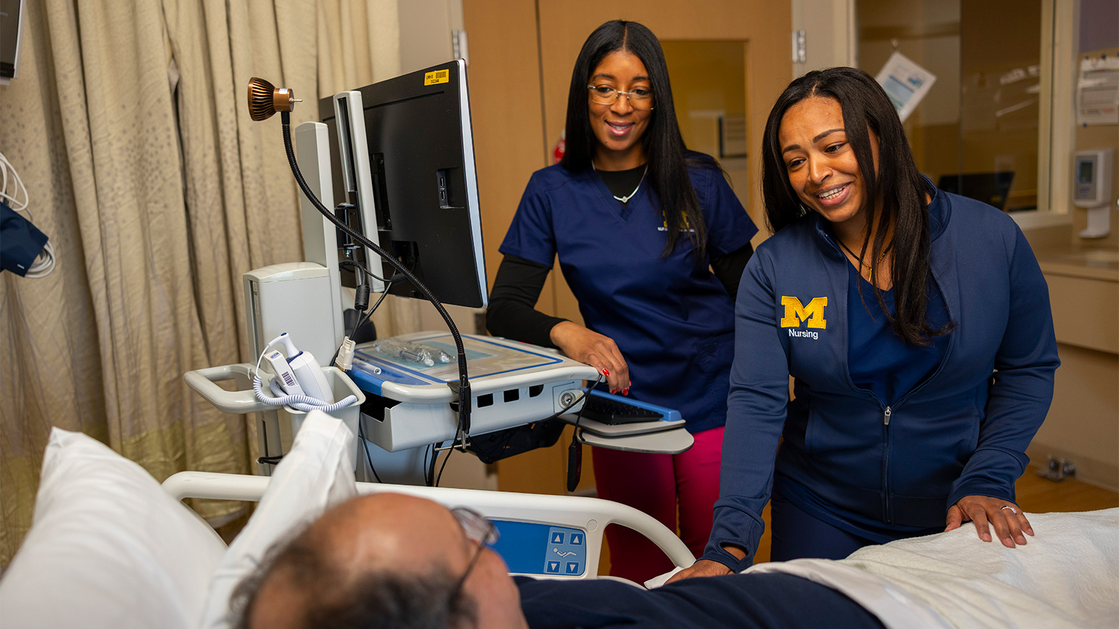 Two U-M nursing professionals smile while interacting with a patient in a hospital room.