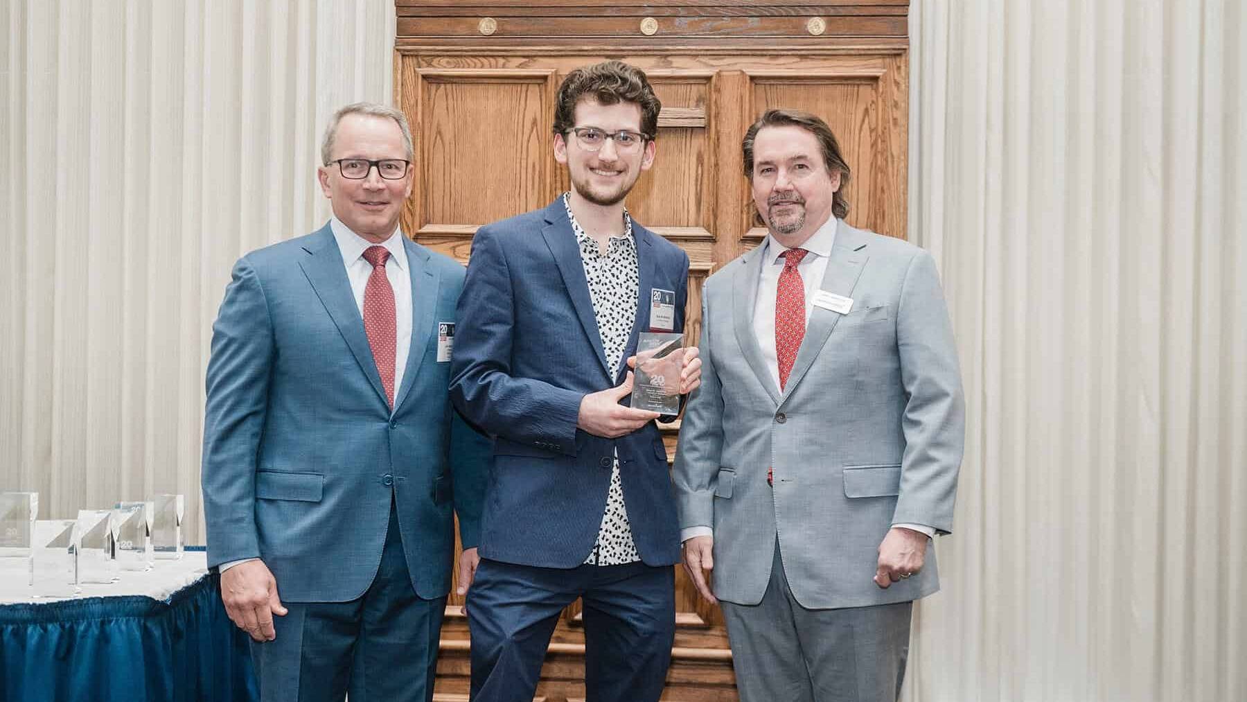 Sinan Abdulhak accepts a glass award while two men stand either side of him. They all wear formal attire.