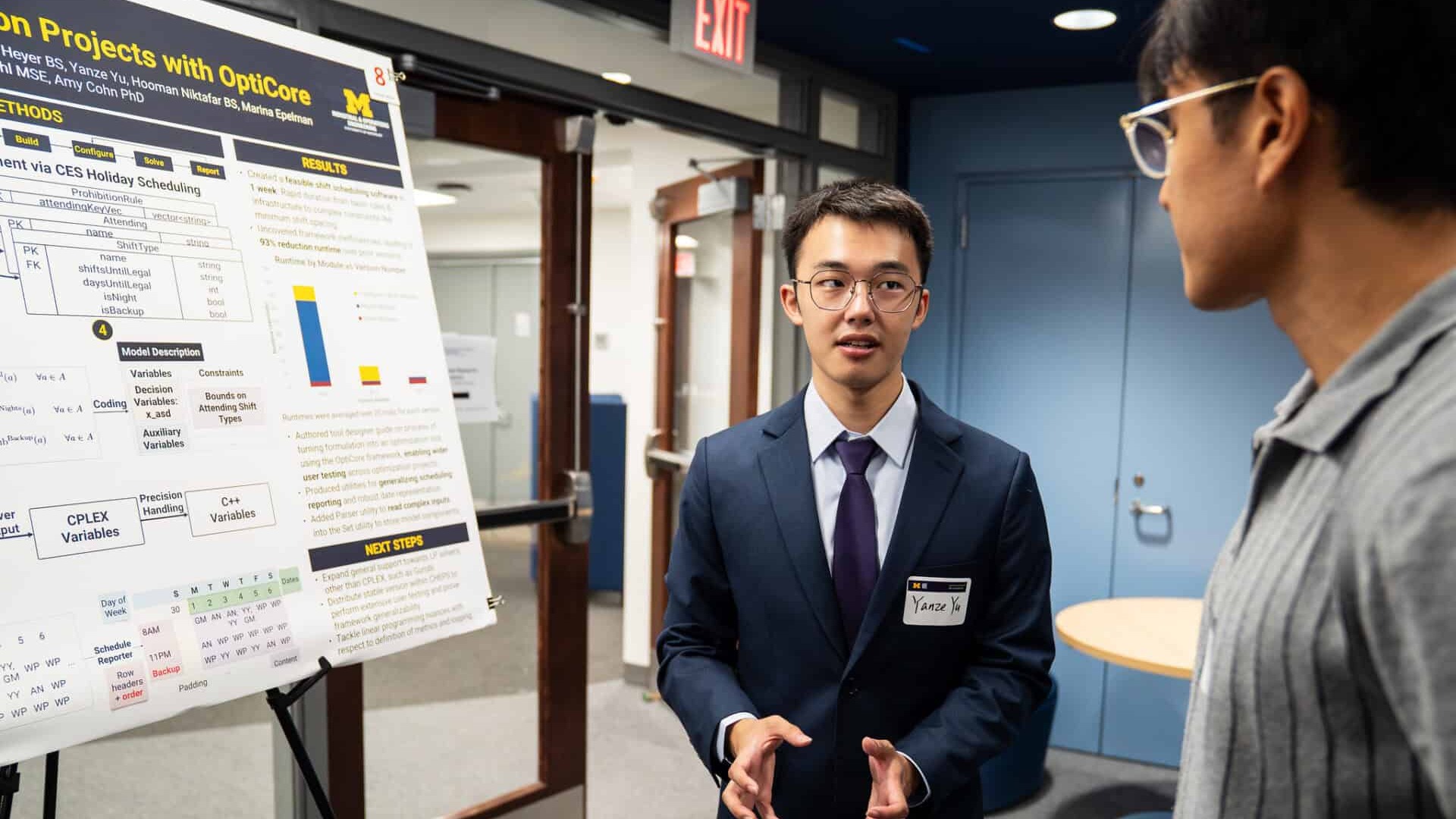 Two people discussing a poster presentation. One individual is gesturing towards the poster while explaining details, and the other is attentively listening.