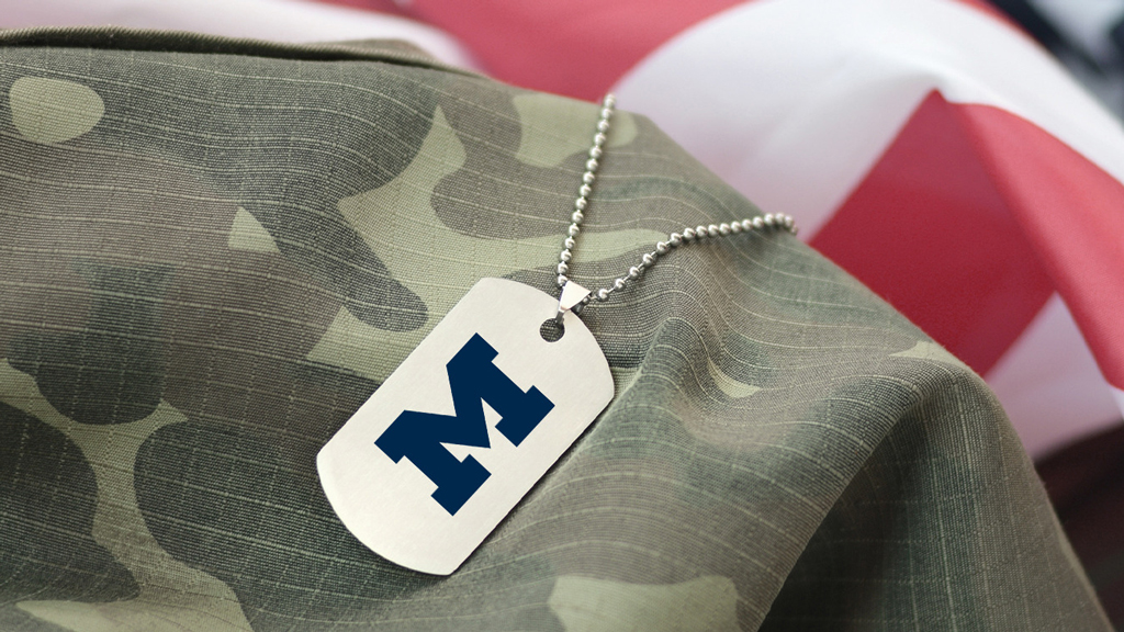 A composite image of a military dog tag with a block M on it draped over a portion of a camouflage jacket with an American flag in the background.