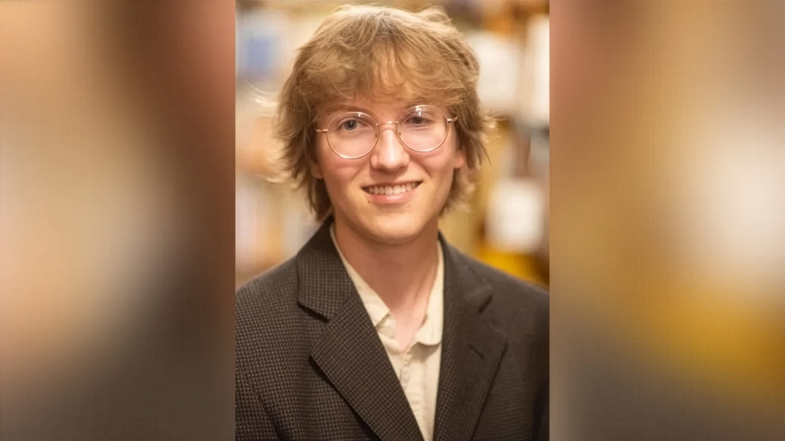 A portrait of Chase, a young person with light brown wavy hair and round wire-rimmed glasses, wearing a light collared shirt under a dark textured blazer and smiling softly.