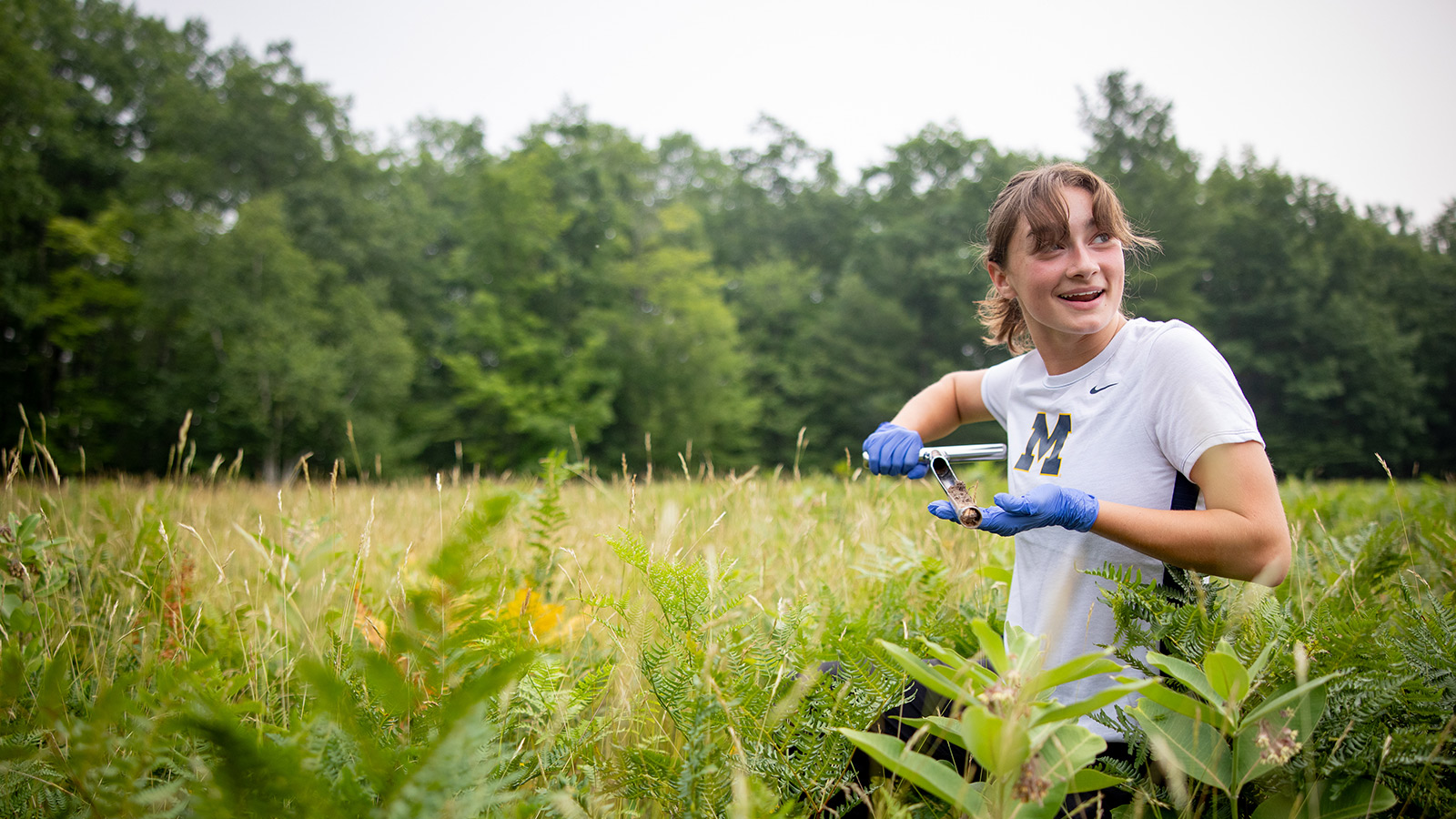 A smiling student wearing a white Michigan shirt and blue gloves holds a metal soil core sampler in a grassy field at the University of Michigan Biological Station.