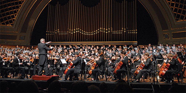 A conductor leads a full orchestra and a large choir in Hill Auditorium, performing on a grand stage in front of an impressive pipe organ.