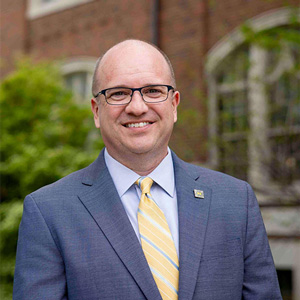 Matt Burrows in a gray suit and yellow striped tie stands outdoors in front of a brick building.