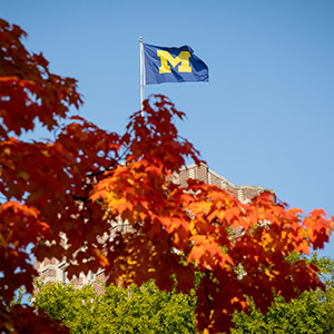 The University of Michigan flag waves atop the Union, rising above vivid red and green autumn leaves beneath a clear blue sky.