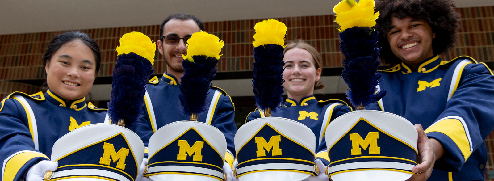 Four band members dressed in Michigan uniforms hold hats with blue and yellow plumes and block M logos.