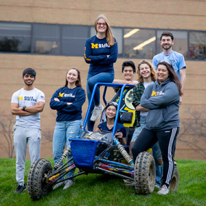 Several students stand and sit around a blue off-road vehicle on the grass in front of a brick building, dressed in Michigan-themed clothing.
