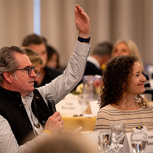 A person holds a microphone and raises a hand while seated at a discussion event.