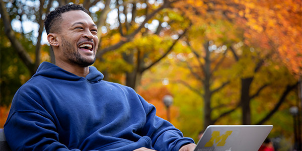 A student sits outdoors with a Michigan laptop, surrounded by fall foliage.
