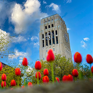 Burton Memorial Tower and its large clock face stand tall against a blue, partly cloudy sky, with red tulips in the foreground and budding spring trees at the base.