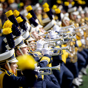 A line of trumpet players dressed in navy and maize uniforms topped with tall blue and yellow plumes, all playing in synchronized formation.