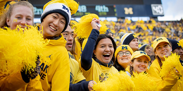 A crowd dressed in yellow and blue waves bright yellow pom-poms, energetically supporting their team at Michigan Stadium, with scoreboard and stands full of fans in the background.