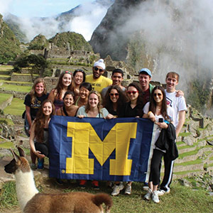 A group poses with a maize block “M” flag in front of terraced stone ruins and lush, misty mountains, while a llama stands nearby.