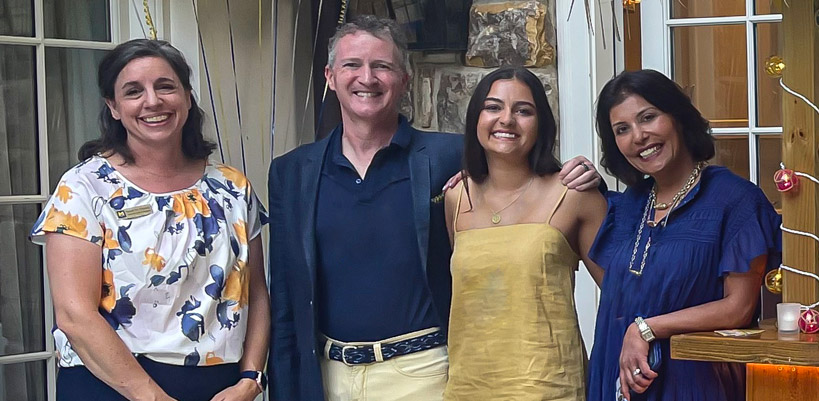 Enu Mainigi and John Walke, standing with their daughter Sonia and another woman, pose together in front of glass doors and a stone wall at a warmly decorated gathering.