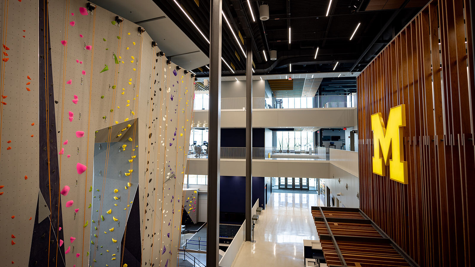 Multi-story Hadley recreation center interior featuring a tall climbing wall with colorful holds and ropes beside a large maize Block M mounted on a wood-paneled wall.