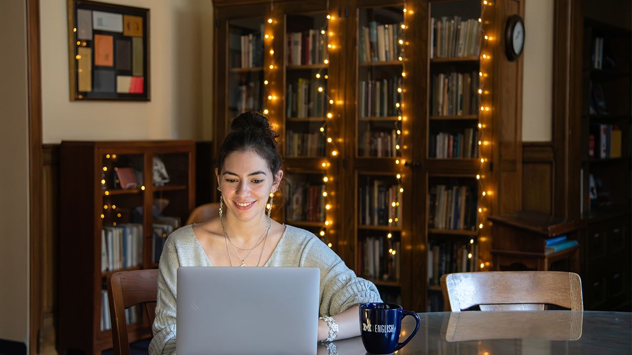 A student works on her laptop computer at a table in a study lounge. In the background are bookshelves decorated with string lights.
