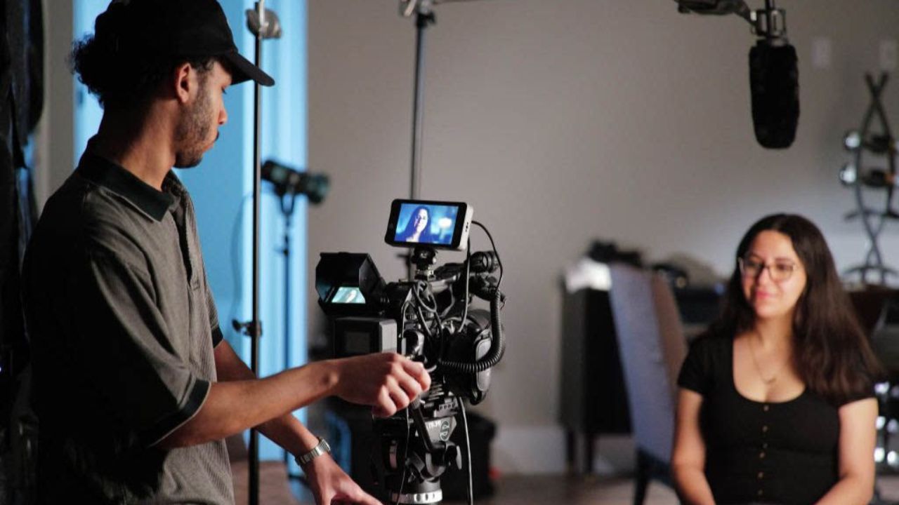 A camera operator films a seated woman in a studio, with a boom microphone suspended overhead.