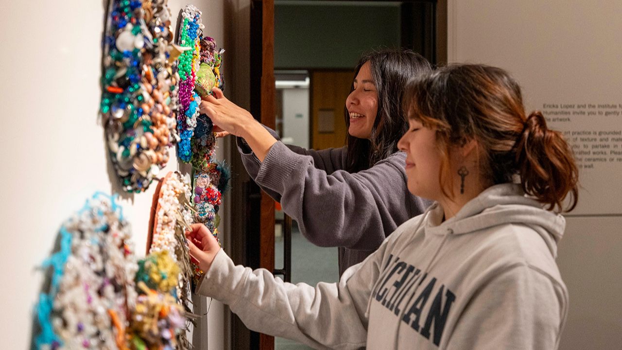Two women touch colorful, textured mixed-media artworks displayed on a gallery wall.