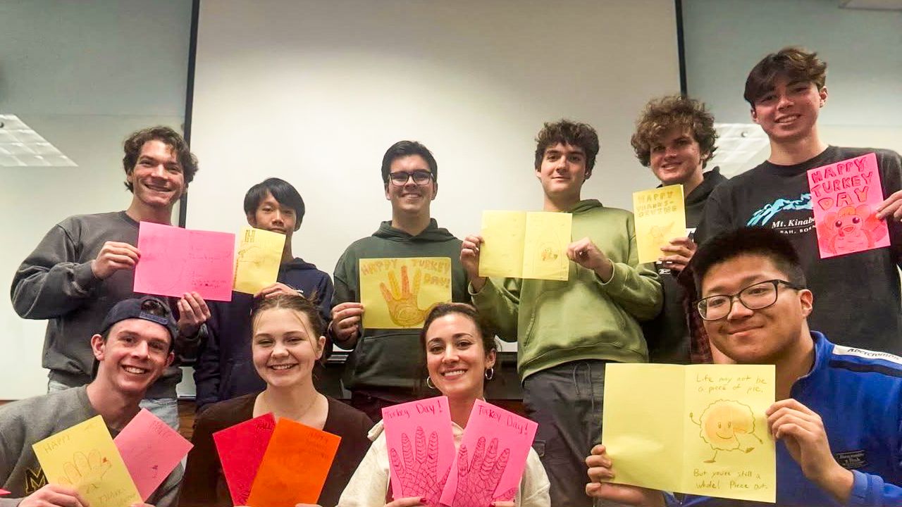 Ten smiling students pose in a classroom while holding colorful handmade Thanksgiving cards.