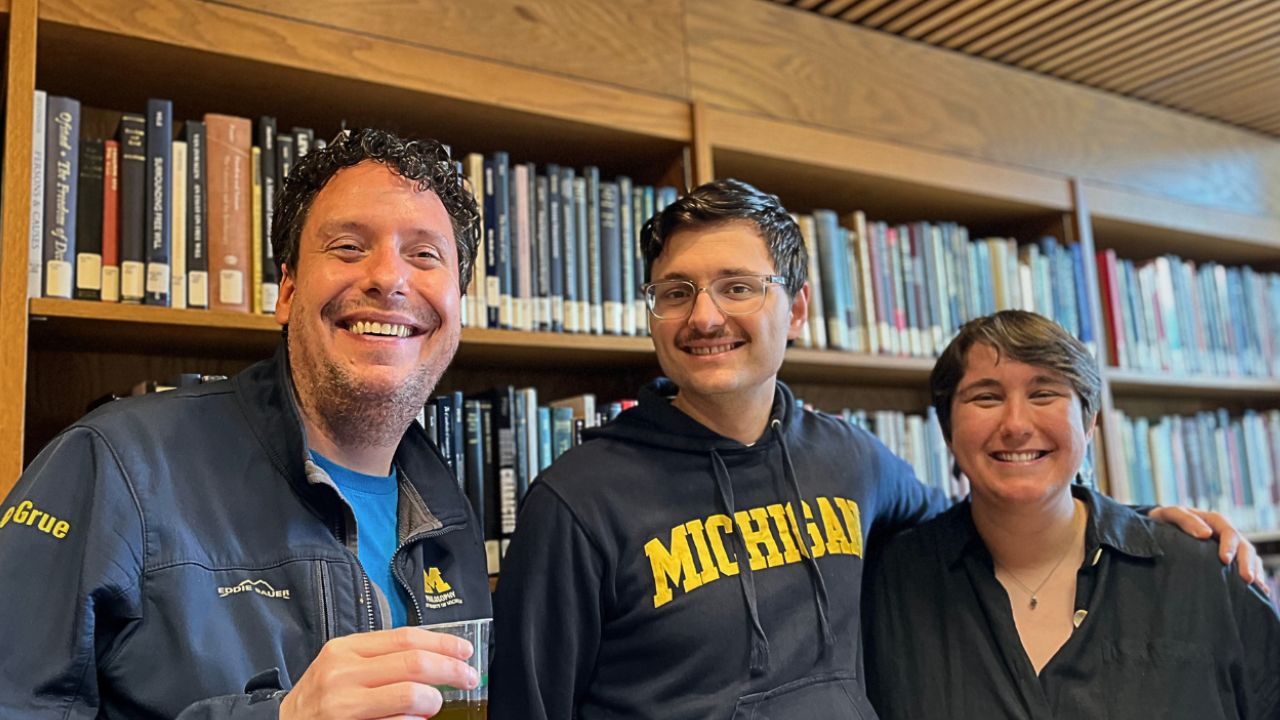Three smiling people pose together in front of library bookshelves; the person in the center wears a Michigan sweatshirt.