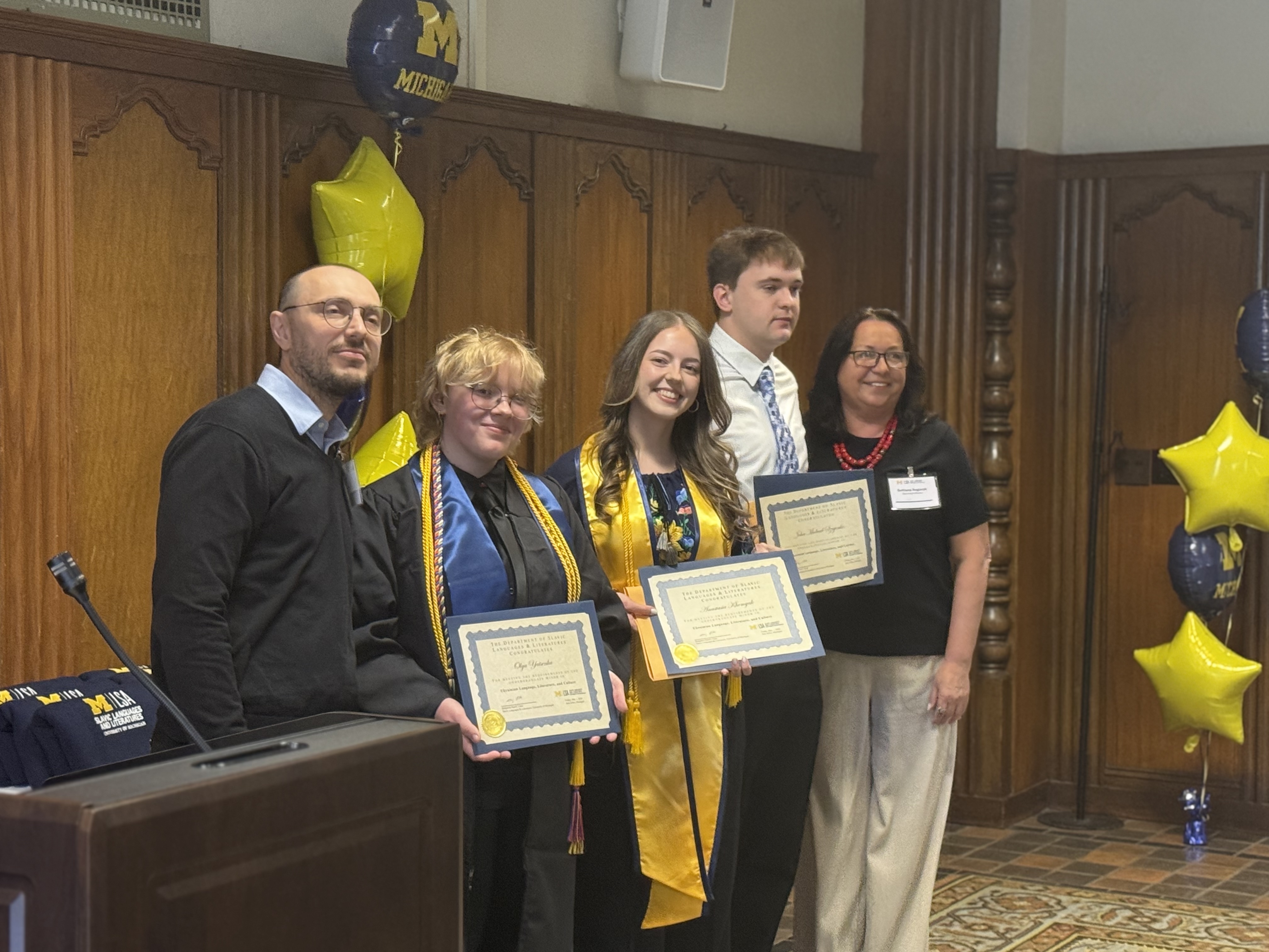 4 people pose together at an indoor celebration beside blue and maize balloons and a podium.