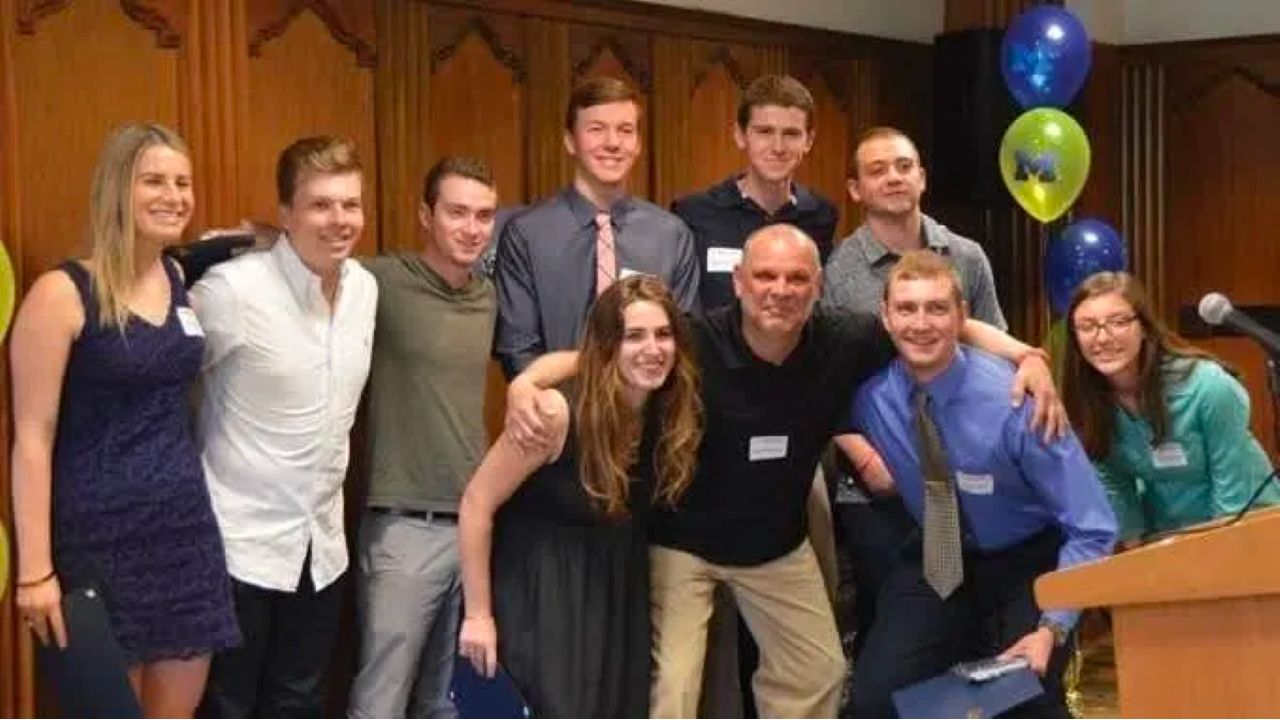 Ten people pose together at an indoor celebration beside blue and maize balloons and a podium.