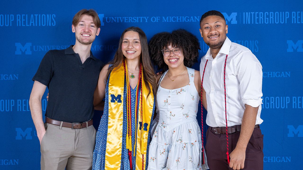 Four smiling University of Michigan students, two wearing graduation stoles or cords, pose before an Intergroup Relations backdrop.